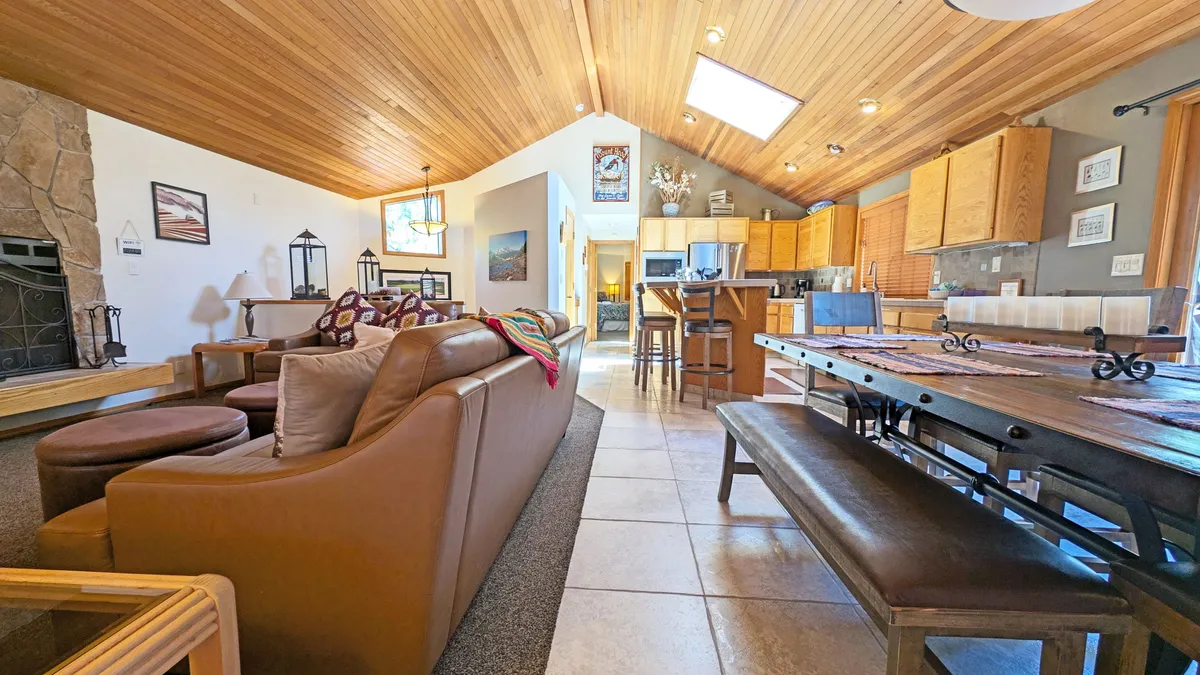 This interior scene centers on a spacious living room featuring a dramatic vaulted wood-paneled ceiling, a substantial stone fireplace, plush leather seating, a cozy throw, and a dining table in the background. Large windows suggest abundant natural light, and tasteful decor accents the warm ambiance.