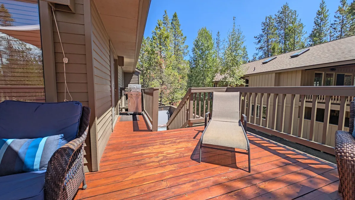The photo presents a spacious wooden deck featuring a lounge chair, barbecue grill, and views of tall trees. The setup invites sunbathing, outdoor dining, and leisure time with friends or family.