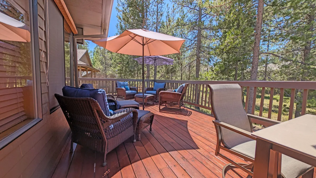 An inviting wooden deck features comfortable outdoor seating under umbrellas and is nestled among tall trees. The ambiance is tranquil, perfect for family meals, relaxing with a book, or enjoying the fresh air together. The deck setting suggests ample space for families to gather, eat, and socialize outdoors.