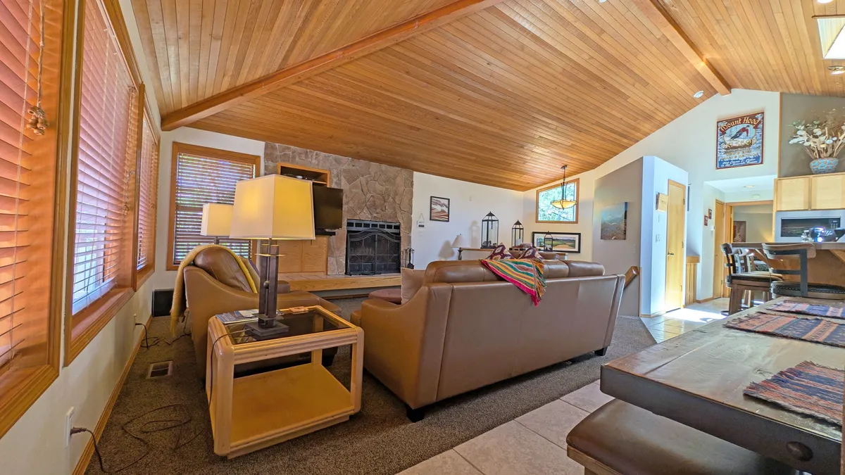A warm, inviting living space with a central fireplace, comfortable sofa, wooden ceiling, and a dining table nearby. Natural light pours in through a large window, enhancing the sense of openness and welcoming guests to relax or socialize.