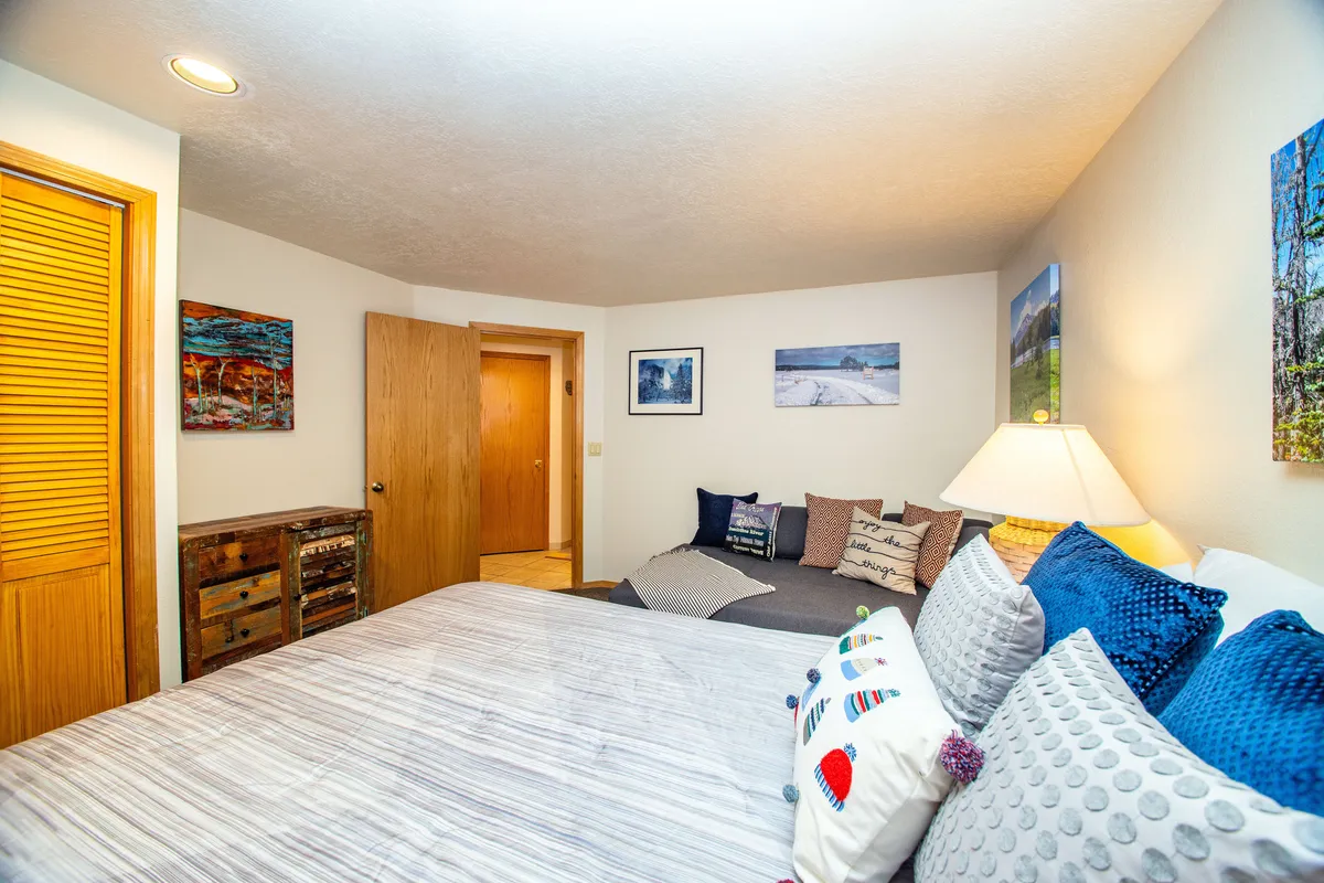 A corner of a bedroom with a bed adorned in decorative pillows, a cozy sofa area, wall-mounted artwork, and a warm lamp. The layout invites quiet relaxation or conversation, with comfortable textures and a welcoming mood.