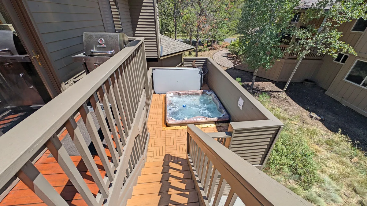 This exterior image features a private hot tub on a deck surrounded by pine trees, with a deck railing and a pathway leading away from the house. The setting suggests a tranquil natural environment ideal for unwinding after outdoor adventures. The hot tub and grill area are visible, inviting guests to relax or barbecue while soaking in the scenery.