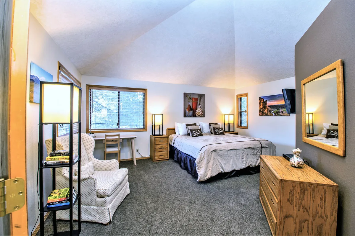This image features a serene bedroom with a king bed, a cozy armchair, a dedicated desk workspace, decorative artwork, and a mirror. Gentle natural light fills the room, and a flat-screen TV is visible, providing a comfortable and functional environment ideal for rest and productivity.