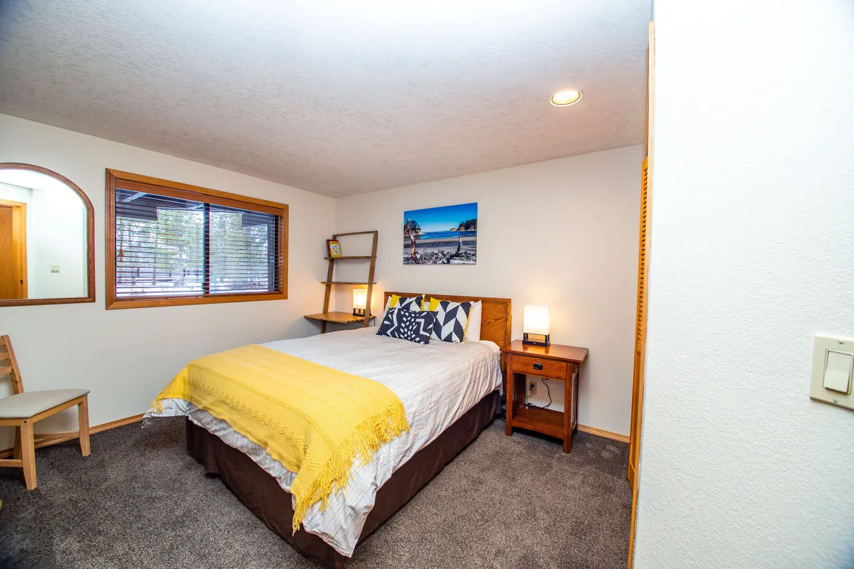 A bright bedroom with a bed adorned with a yellow throw, a nightstand with a lamp, cheerful wall art, and a window with blinds. The space is illuminated by natural light, creating a welcoming atmosphere for rest or reading.