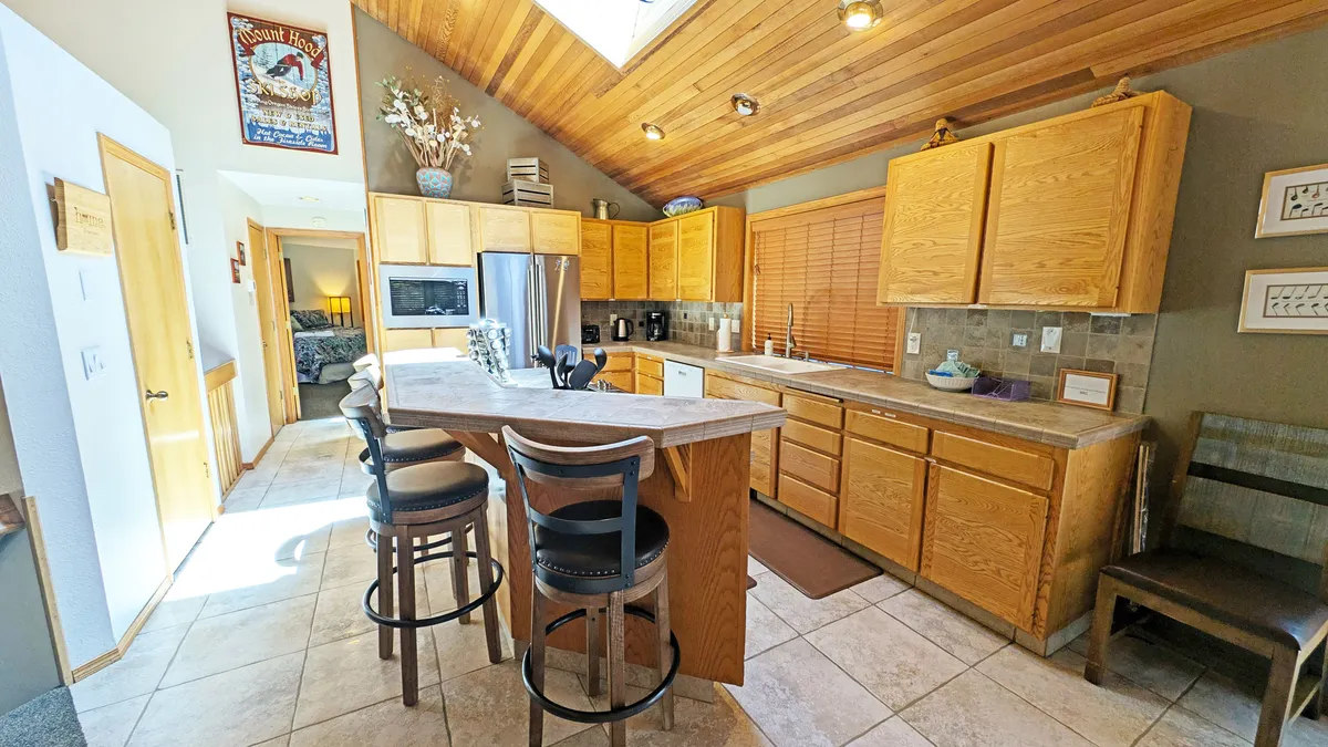 This image features a welcoming kitchen scene with a central island, sleek wooden cabinets, skylight flooding the space with light, and bar stools inviting guests to sit. The layout suggests ample space for multiple cooks, socializing, and group meal preparation, perfectly fitting the description of a well-equipped kitchen designed for gatherings.