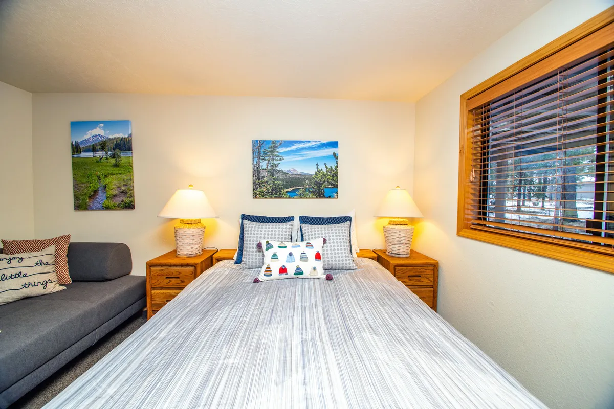 This image captures a bedroom with striped bedding, a cushioned sofa, decorative throw pillows, and wall art featuring scenic landscapes. Natural light streams in through the window, and the inviting seating area is positioned to enjoy views outside.