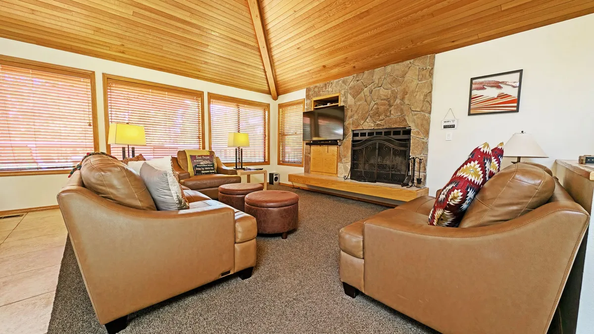 The image showcases a spacious, sunlit living room with a striking wood-burning fireplace as the centerpiece, surrounded by comfortable seating. The high wooden ceilings and expansive windows let in natural light, creating a cozy and inviting space ideal for family gatherings or relaxing after a day of exploring Sunriver.