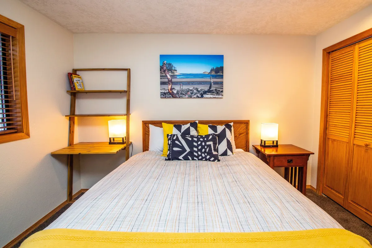 This inviting bedroom features a comfortable bed accented with colorful pillows, calming wall art, and a wooden shelving unit. The setting is bright and peaceful, making it ideal for relaxation or a quiet retreat after a day of activities.